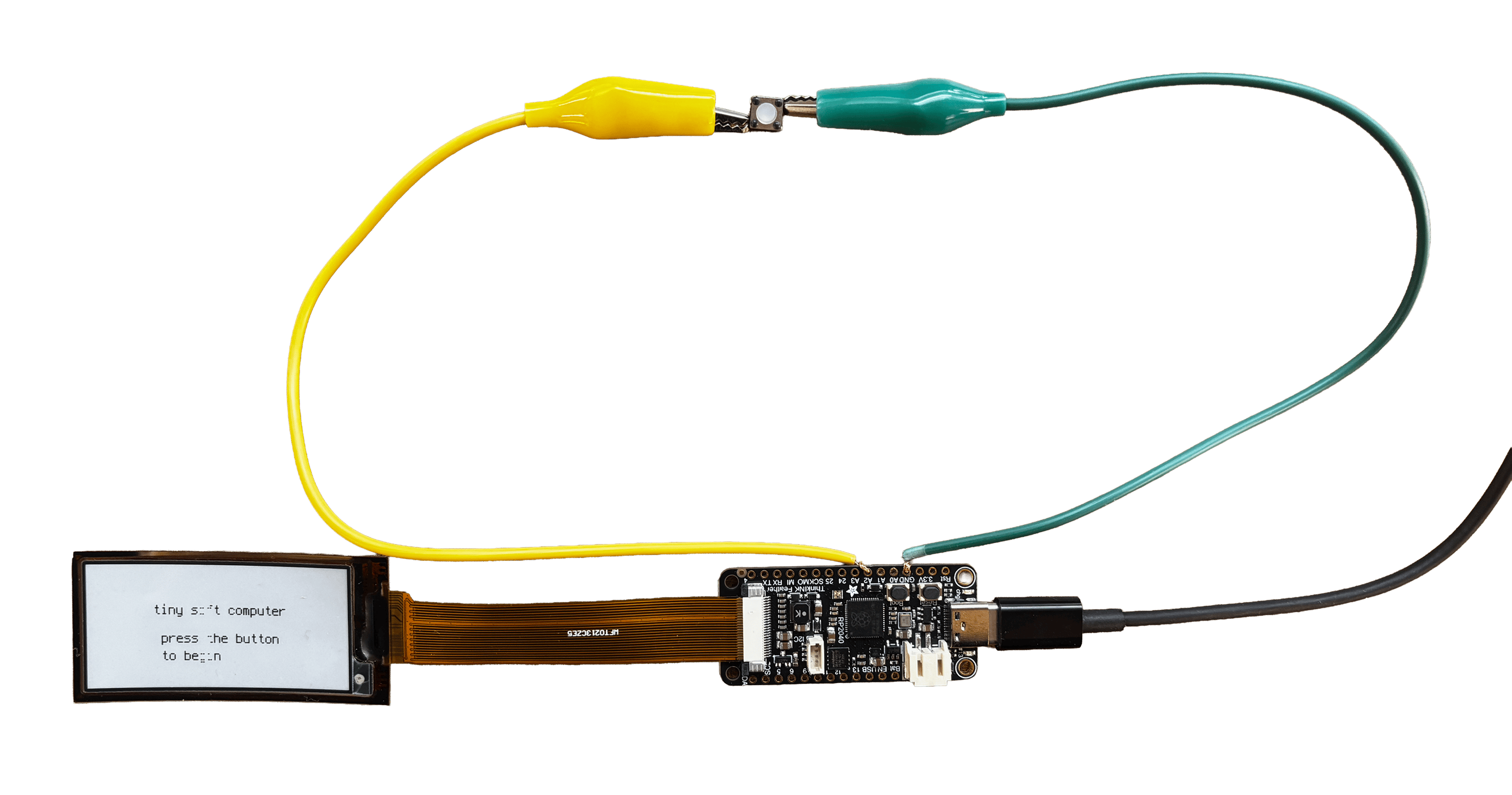 A fully wired tiny soft computer: display connected and showing 'tiny soft computer, press the button to begin', button wired via yellow and green alligator clips to the A2 and GND pins, USB-C cable attached.